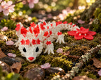 Roze met rode 3D-geprinte cherryblossom baby draak liggend in een natuurlijke bosomgeving met mos en bloesem.