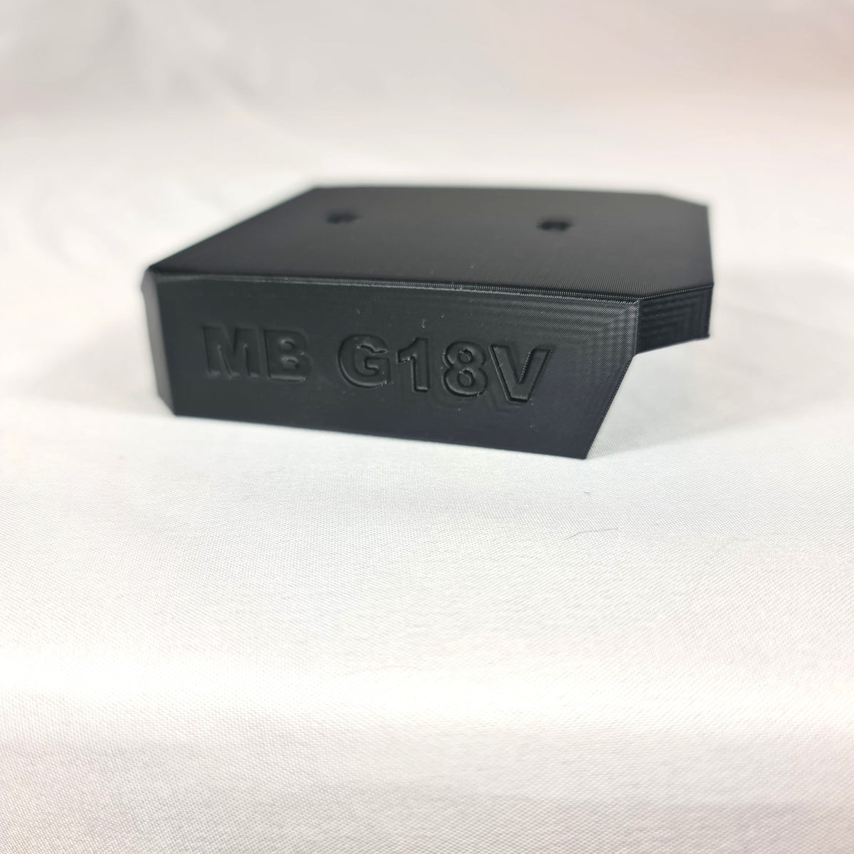 Close-up opname van de losse 3D-geprinte zwarte wandhouder en opbergbeugel voor Makita G-Series 18V accu's op witte stof
